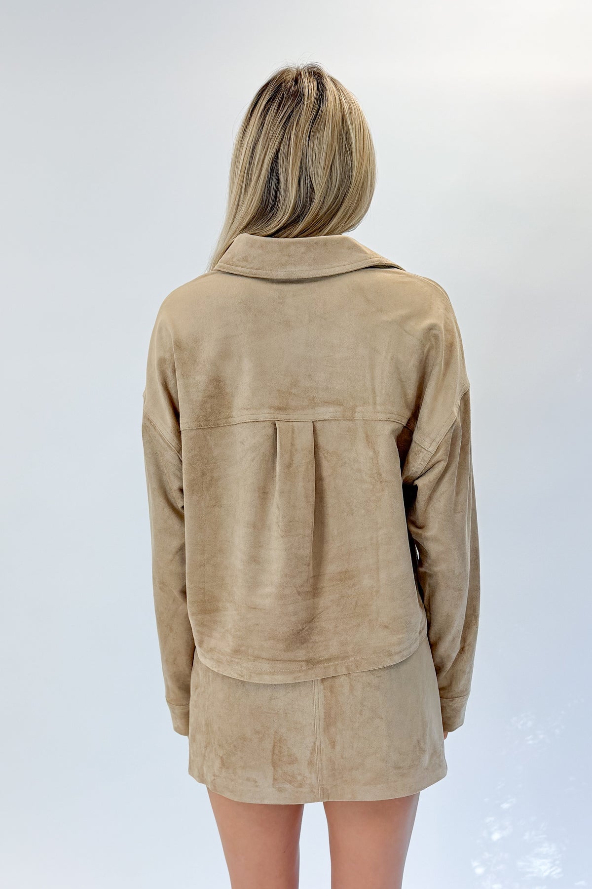 Back view of woman wearing Swagger Boutique’s Light Sand All Day Cropped Suede Jacket and mini skirt by Z Supply against white studio background.