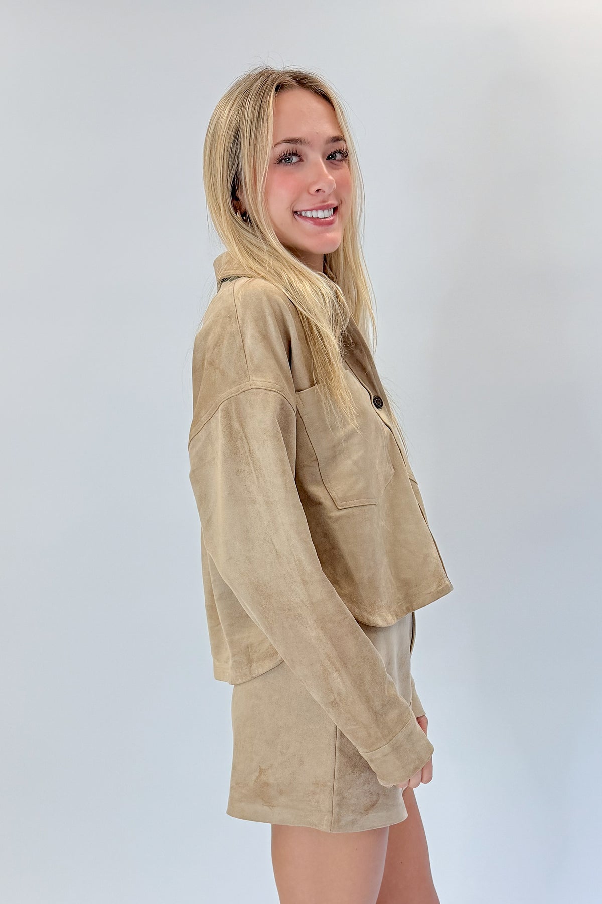 Side view of woman wearing Swagger Boutique’s Light Sand All Day Cropped Suede Jacket and mini skirt by Z Supply against white studio background.