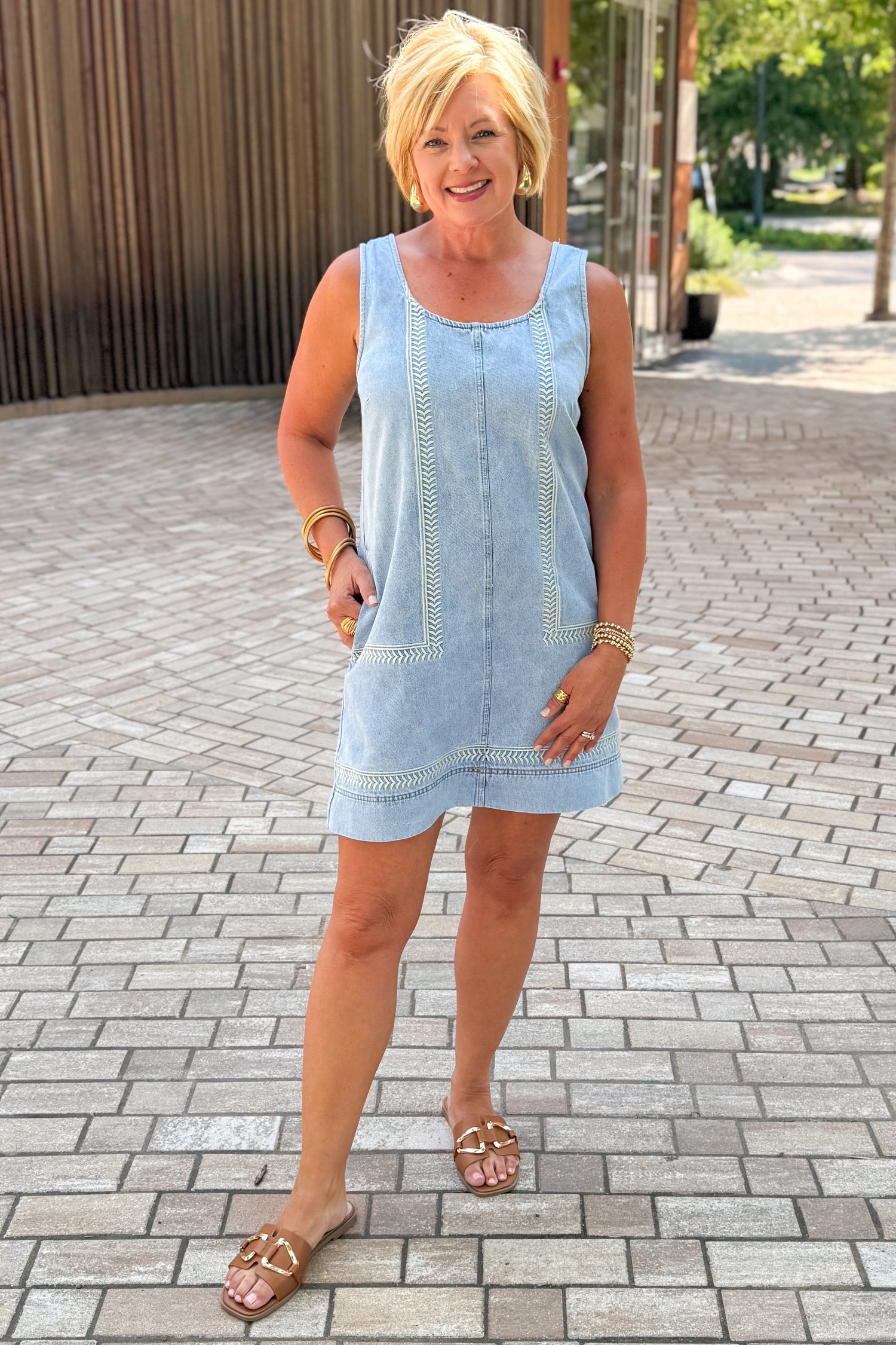 Front view of woman wearing Swagger Boutique’s Light Wash Embroidered Pocket Mini Dress with sandals standing outside at a park.
