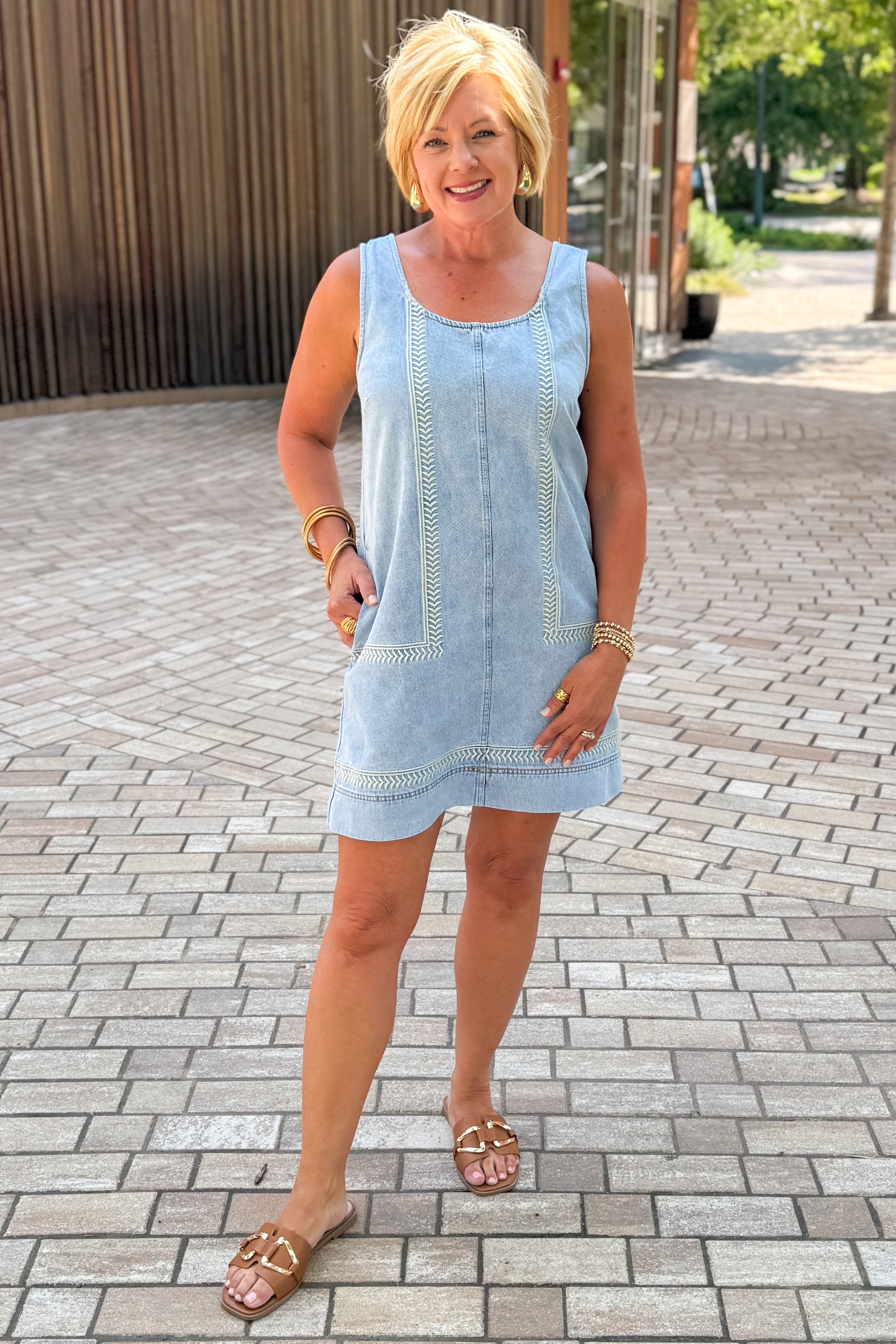 Front view of woman wearing Swagger Boutique’s Light Wash Embroidered Pocket Mini Dress with sandals standing outside at a park.