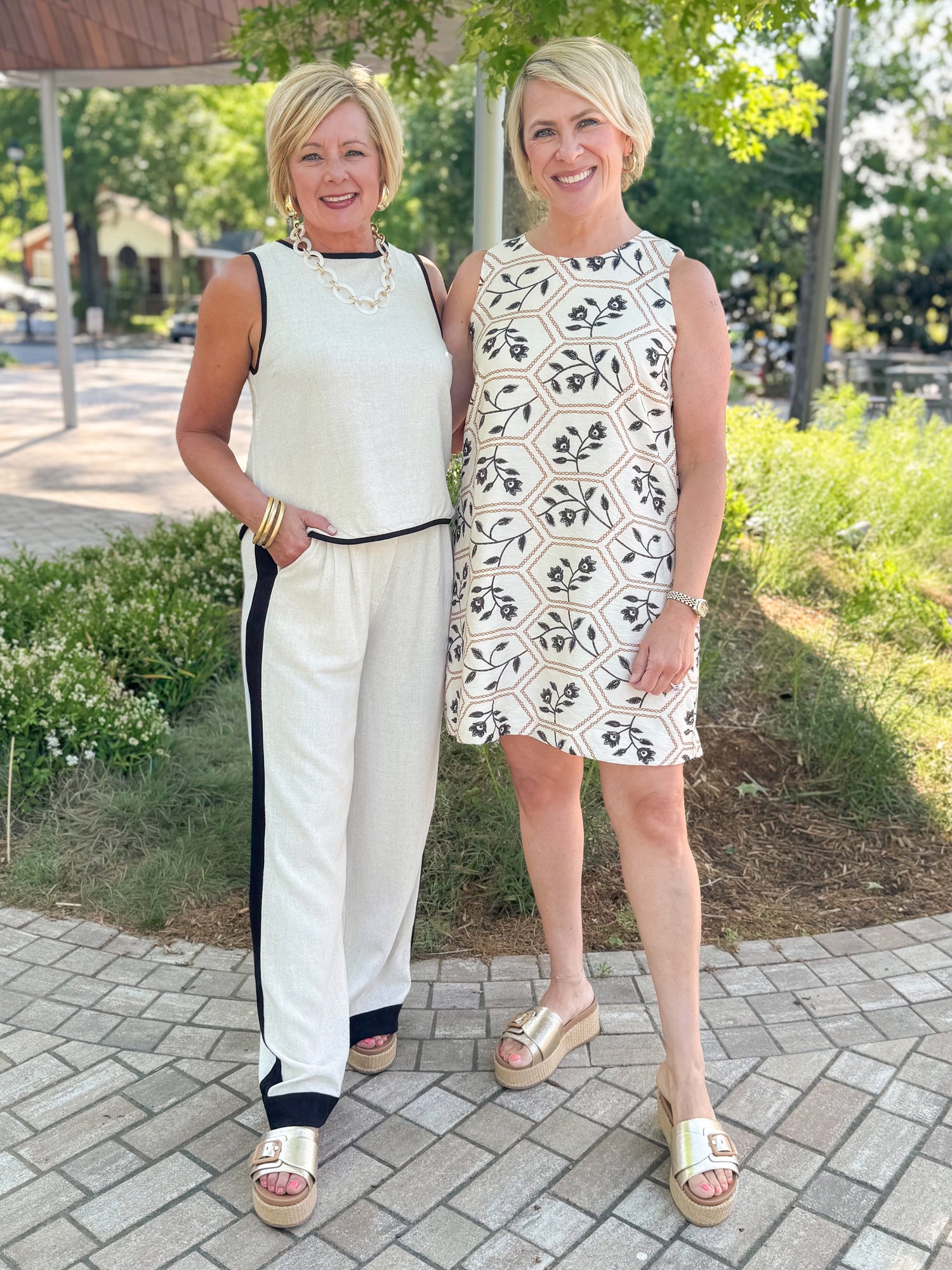 Front view of woman wearing Swagger Boutique’s Linen &amp; Black Lined Tank with matching Linen &amp; Black Stripe Pants and platform sandals with another woman wearing Cream, Taupe &amp; Black Floral Printed Shift Dress outside at a park.