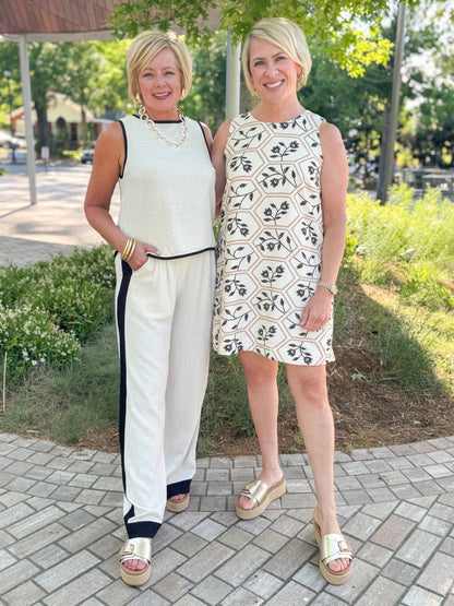 Front view of woman wearing Swagger Boutique’s Linen &amp; Black Lined Tank with matching Linen &amp; Black Stripe Pants and platform sandals with another woman wearing Cream, Taupe &amp; Black Floral Printed Shift Dress outside at a park.