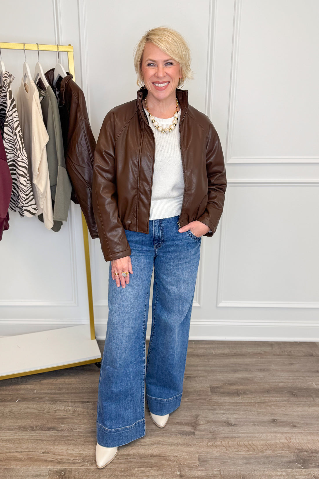 A woman stands indoors wearing a brown leather jacket, white top, wide-leg blue jeans, and white pointed shoes. A clothing rack with other garments is visible on the left, and the background is a white paneled wall with wood flooring.