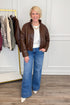 A woman stands indoors wearing a brown leather jacket, white top, wide-leg blue jeans, and white pointed shoes. A clothing rack with other garments is visible on the left, and the background is a white paneled wall with wood flooring.