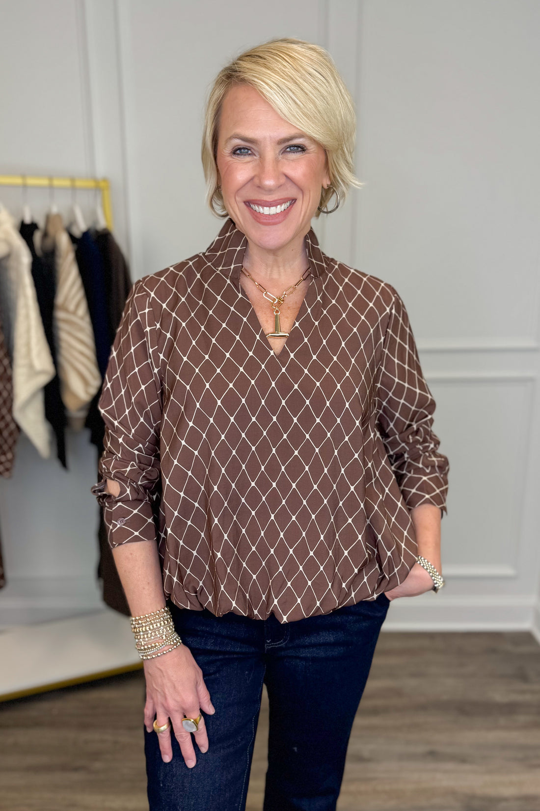 A smiling blonde woman wearing a brown long-sleeve blouse with a white geometric diamond pattern, paired with dark blue jeans, standing in a boutique setting.