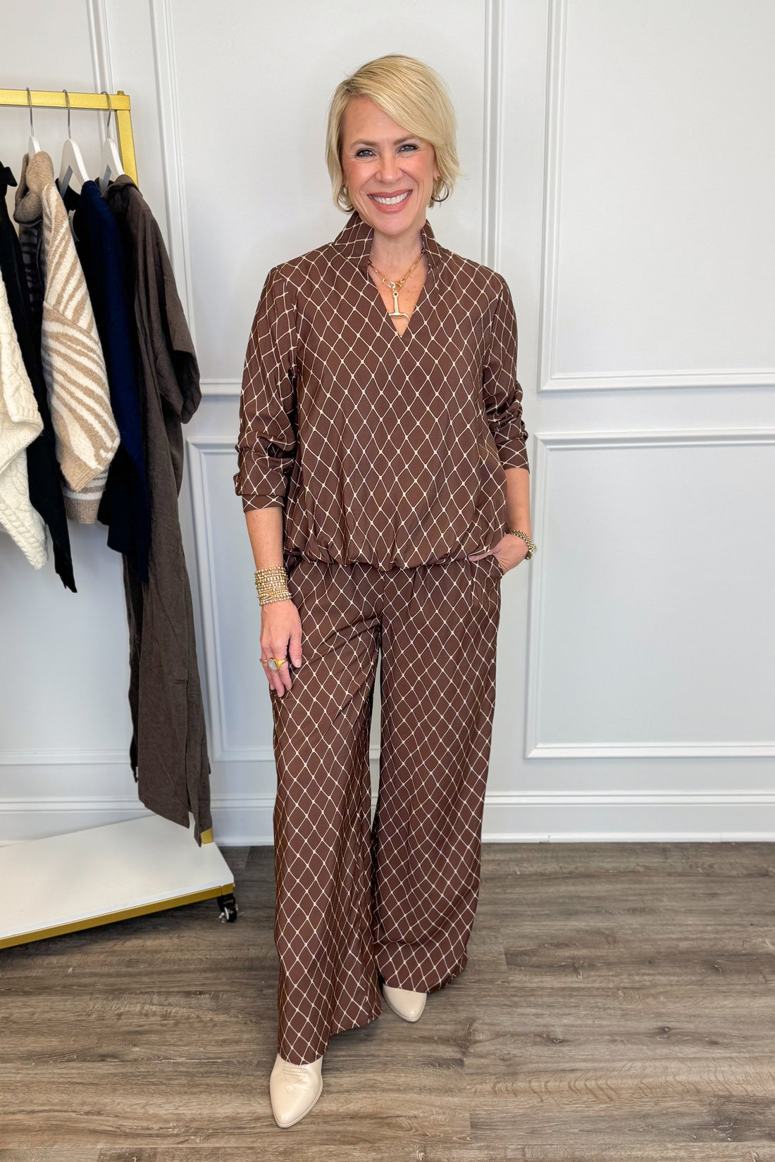 A woman modeling a brown and beige geometric patterned two-piece set consisting of a collared long-sleeve top and matching wide-leg pants, standing in a boutique setting.