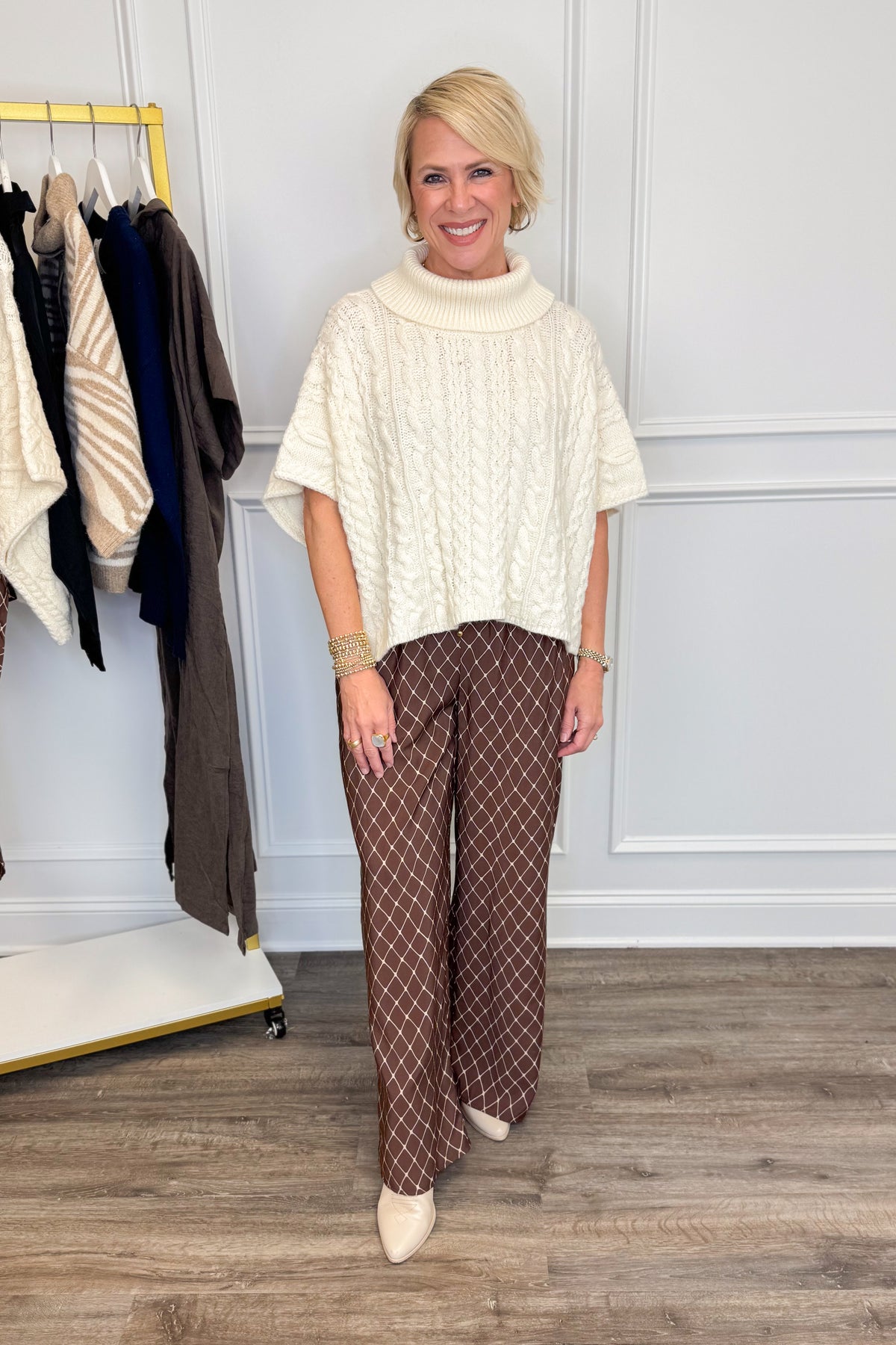 A woman modeling a cream cable-knit mock-neck tunic top and brown wide-leg trousers with a white geometric pattern, standing in a room with a clothing rack and wood floor.