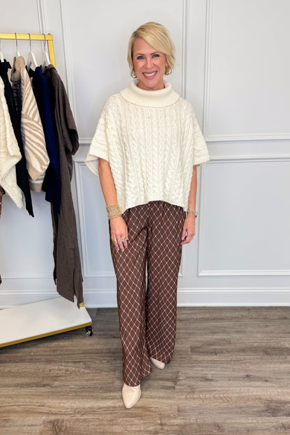 A woman modeling a cream cable-knit mock-neck tunic top and brown wide-leg trousers with a white geometric pattern, standing in a room with a clothing rack and wood floor.