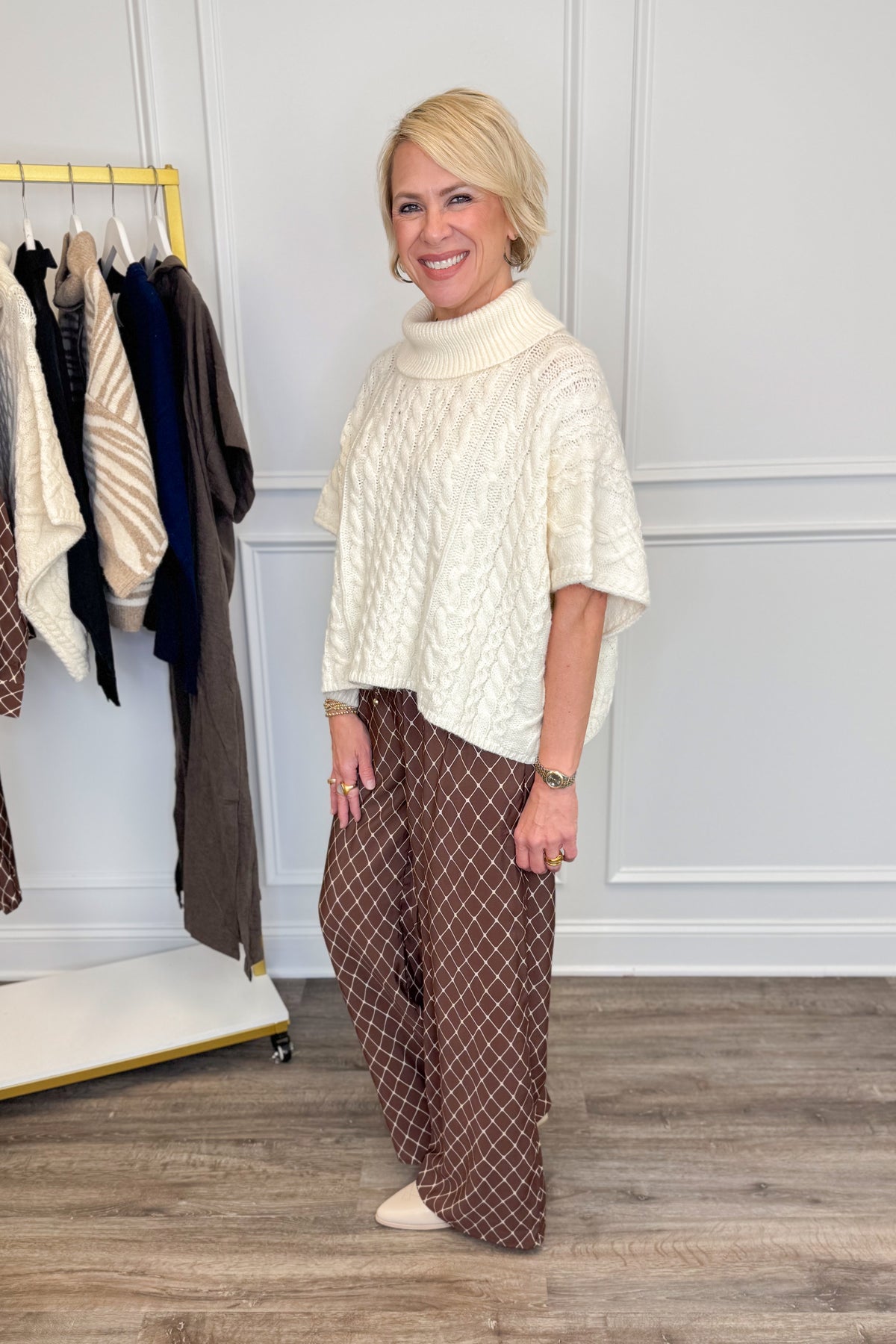 A woman modeling a cream cable-knit mock-neck tunic top with short sleeves, paired with brown wide-leg pants featuring a white geometric pattern, standing in a boutique setting.