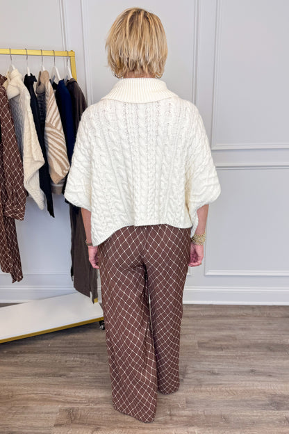 A woman viewed from behind wearing a cream cable-knit cropped cardigan and brown wide-leg trousers with a white grid pattern, standing in a room with a clothing rack and wood floor.