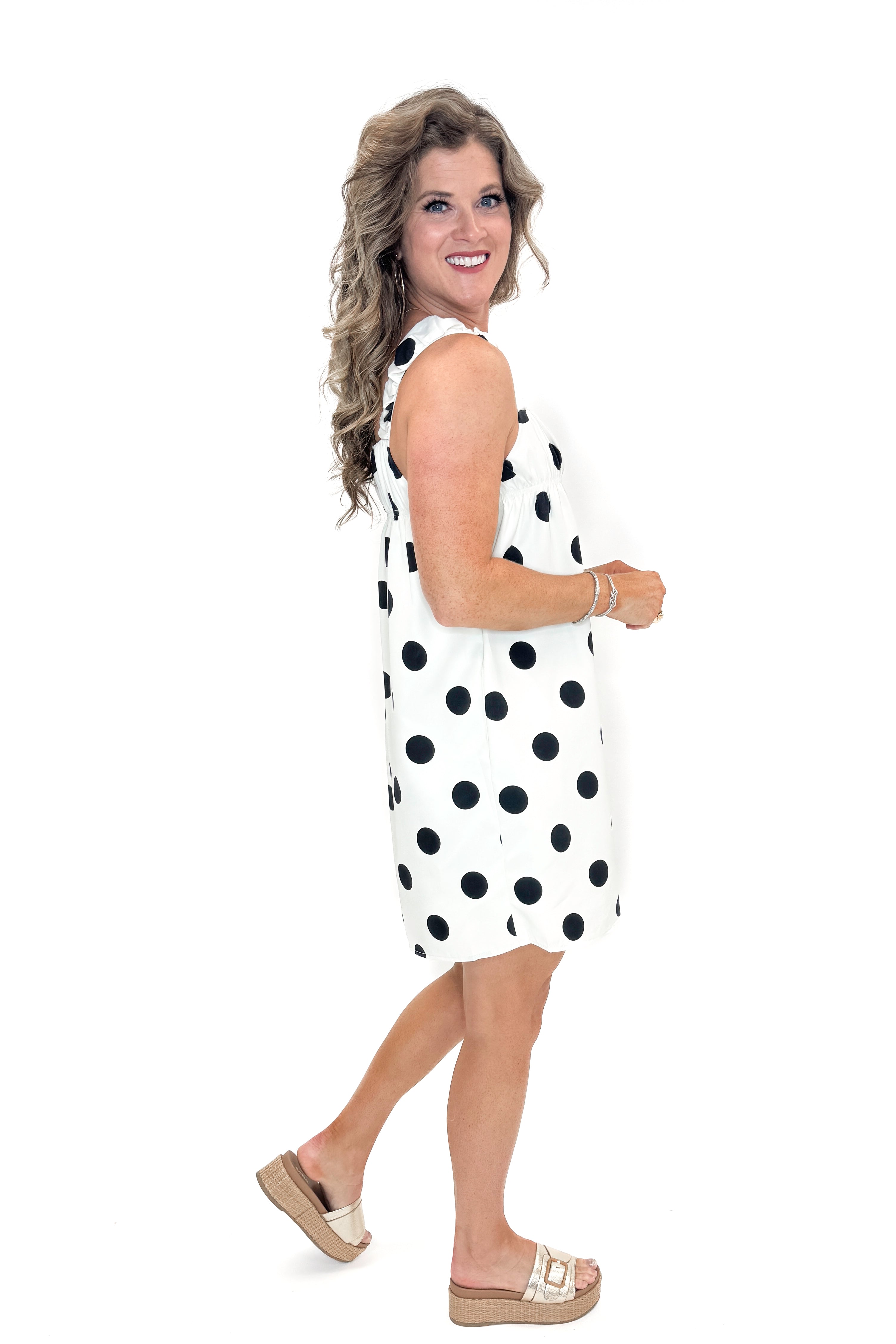 Side view of woman wearing Cream &amp; Black Polka Dot Tank Dress with sandals against white studio background.