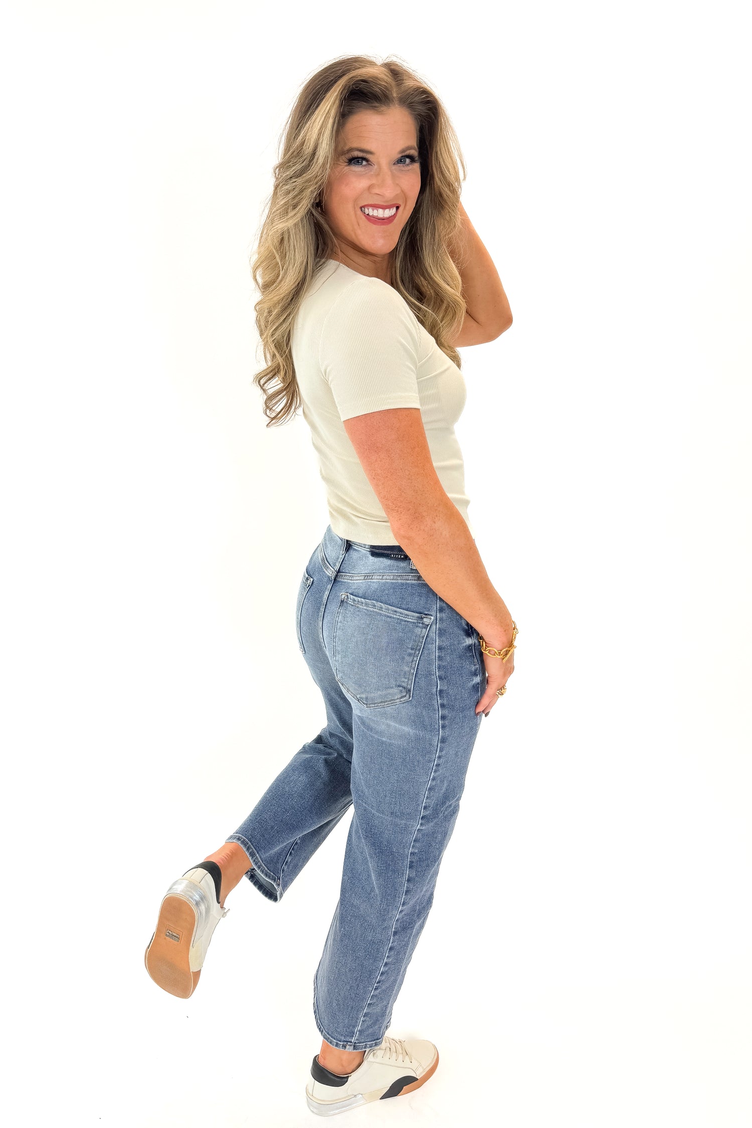 Side view of woman wearing Cream Martini Beaded Ribbed Top with jeans against white studio background.