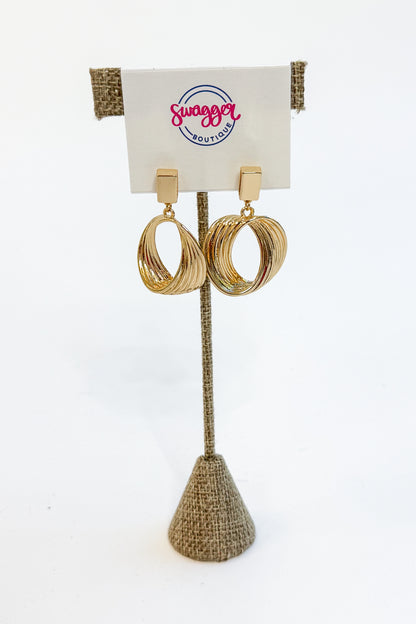 A pair of gold-toned triple-hoop dangle earrings displayed on a burlap-covered stand with a white card labeled &