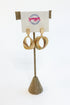 A pair of gold-toned triple-hoop dangle earrings displayed on a burlap-covered stand with a white card labeled &