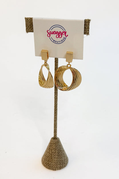 A pair of gold-toned statement earrings displayed on a burlap-covered stand with a white card labeled &