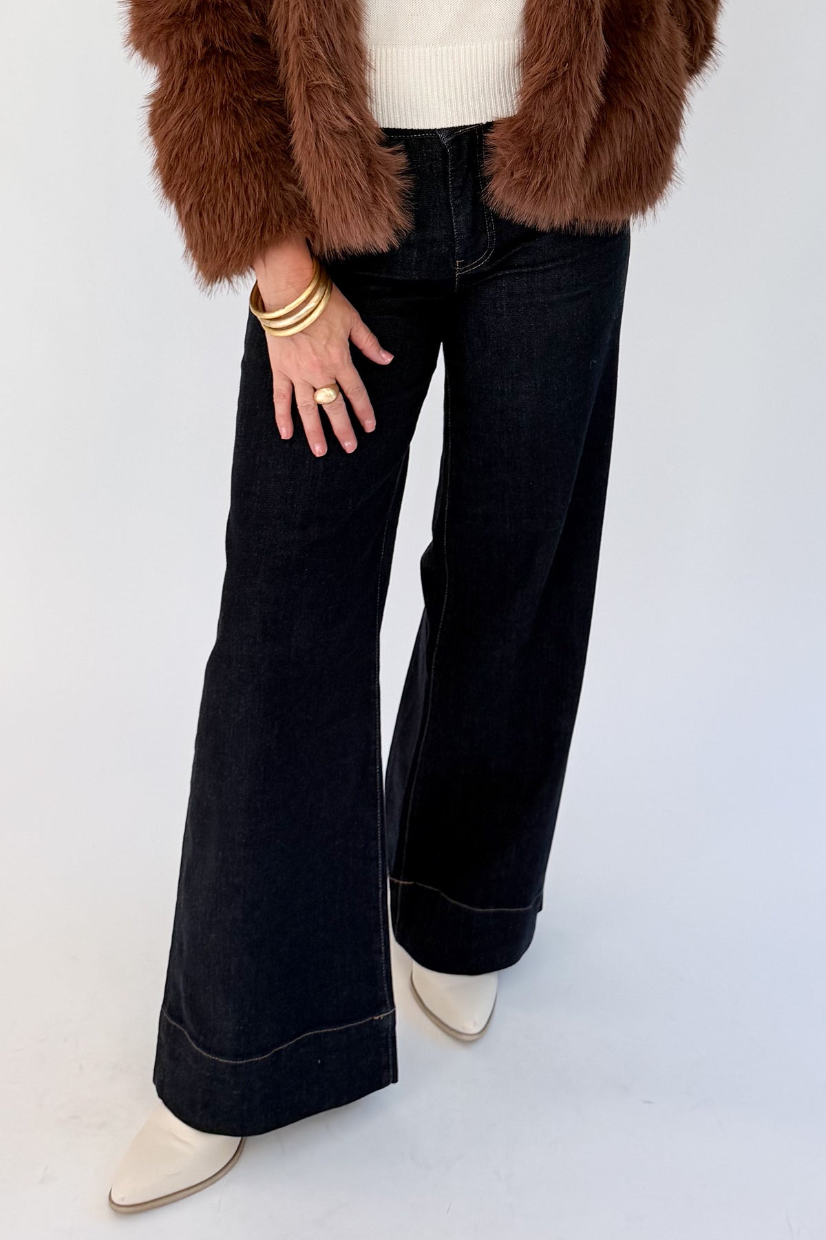 A person wearing dark wide-leg denim jeans with rolled cuffs, styled with a brown faux-fur jacket, cream sweater, cream pointed-toe boots, and gold bracelets and rings, against a plain light gray studio background.