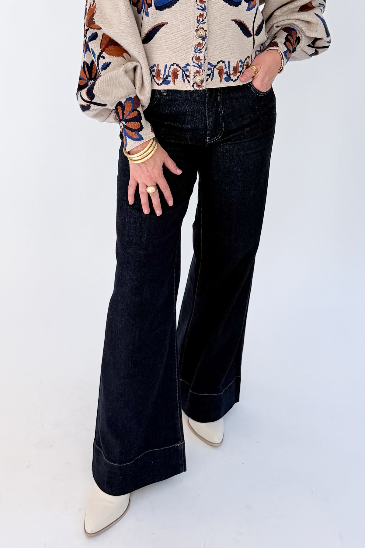 A person wearing dark wide-leg flare jeans with contrast light stitching, paired with a beige patterned blouse and white pointed-toe heels, standing against a plain white studio background.