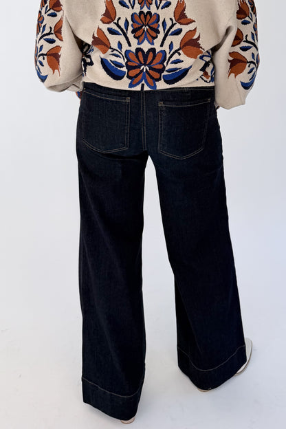 A person shown from the back wearing dark wide-leg denim jeans and a cream blouse with a colorful floral pattern, photographed against a plain light background.