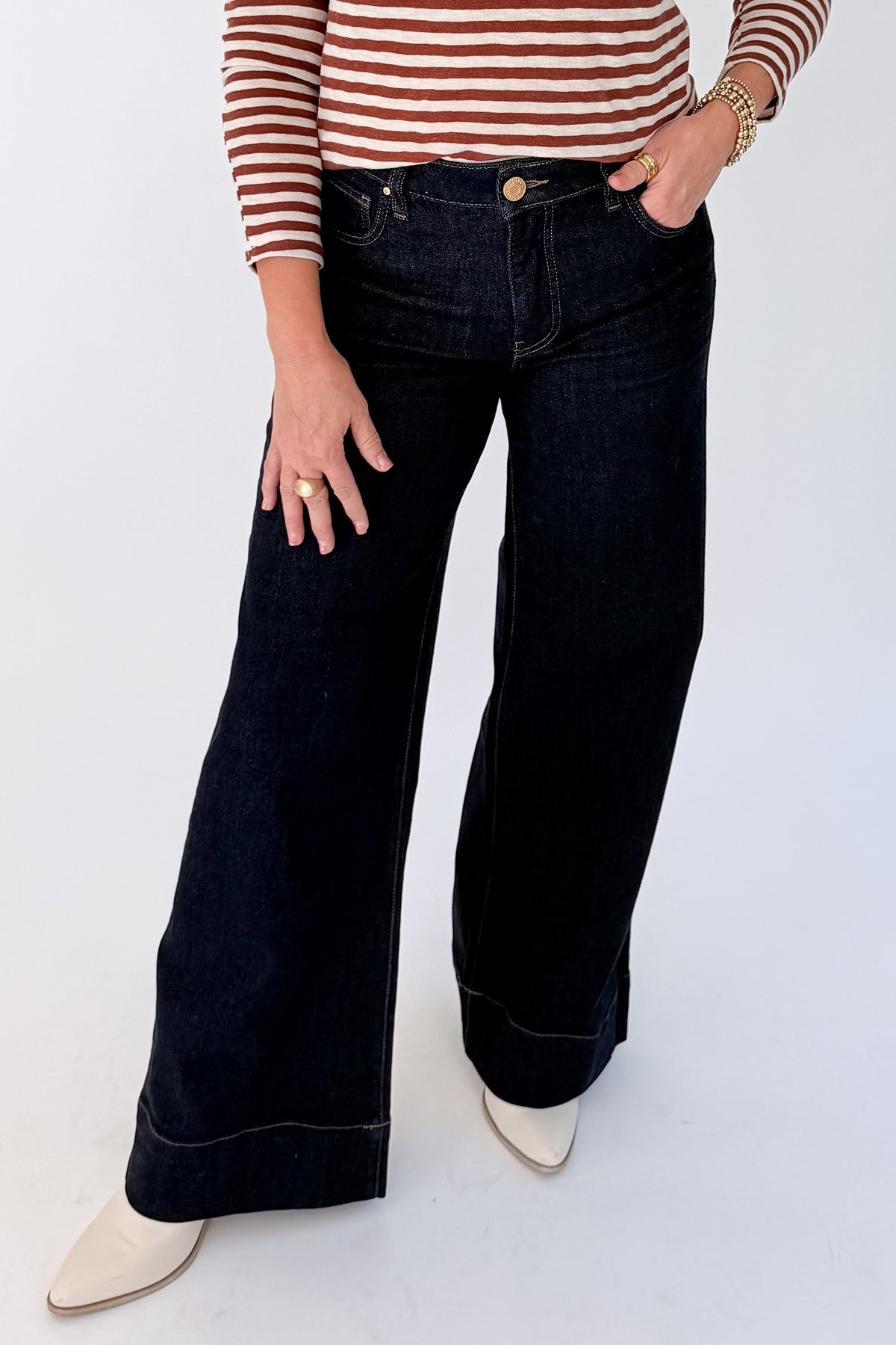 A woman shown wearing a brown and white horizontally striped long-sleeve top, dark wash wide-leg pants, and cream-colored shoes, standing against a plain white studio background.