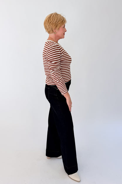 A woman shown in side profile wearing a brown and white horizontally striped long-sleeve top, dark wash wide-leg pants, and cream-colored shoes, standing against a plain white studio background.