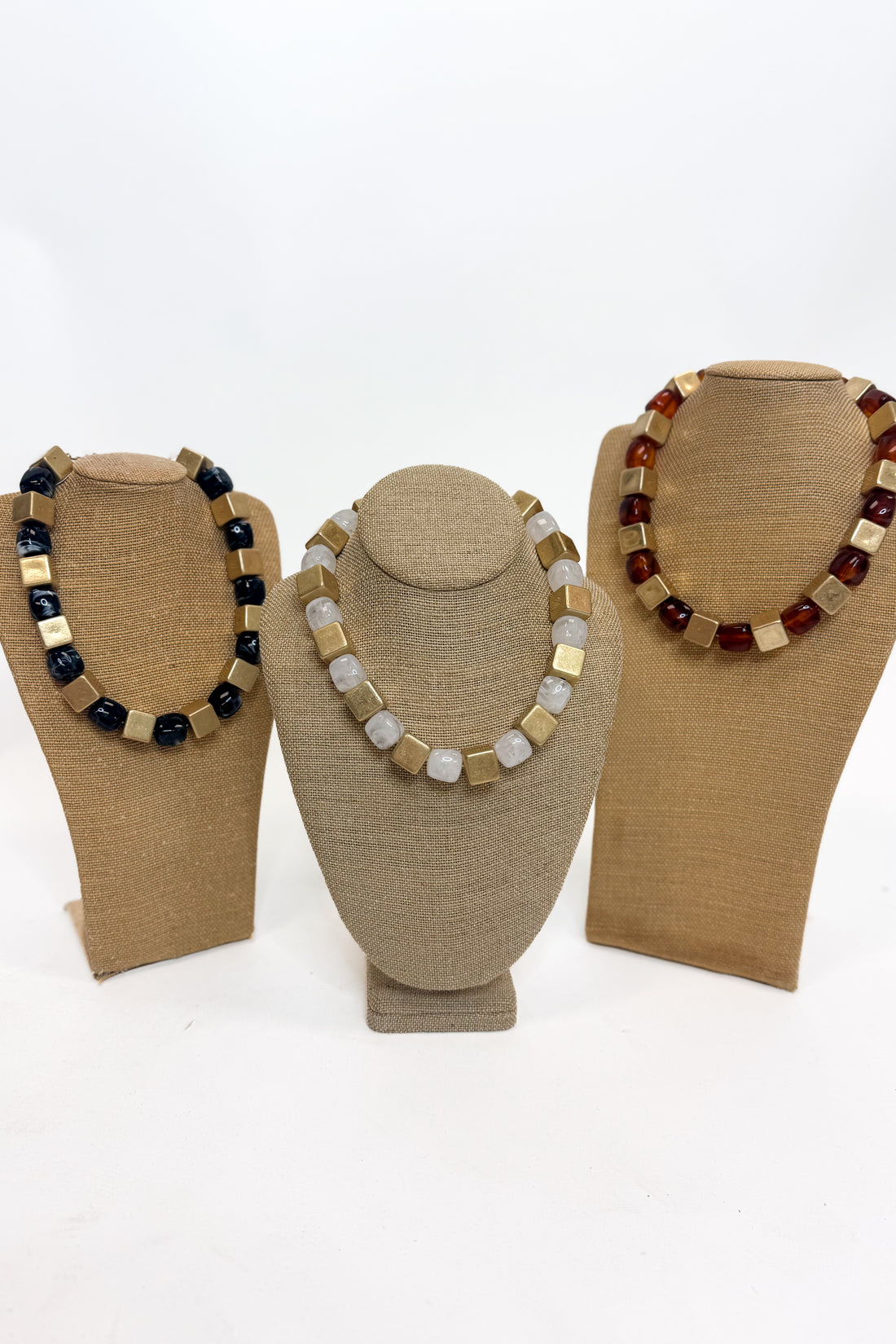 Three chunky beaded necklaces of  alternating polished gold cube beads and stone beads, displayed on tan burlap-covered jewelry busts against a white background.
