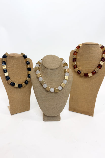Three chunky beaded necklaces of  alternating polished gold cube beads and stone beads, displayed on tan burlap-covered jewelry busts against a white background.