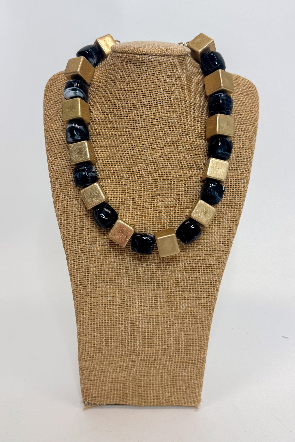 A chunky beaded necklace with alternating polished gold cube beads and black stone beads, displayed on a tan burlap-covered jewelry bust against a white background.