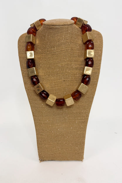 Chunky statement necklace with alternating translucent amber-brown beads and square gold-tone metal cubes, displayed on a tan fabric jewelry bust against a white background.