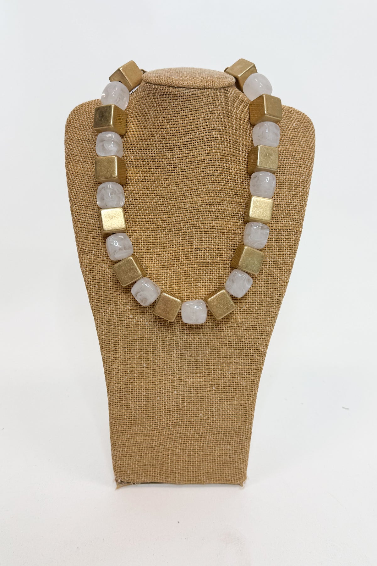 A chunky beaded necklace with alternating matte gold square beads and translucent white-gray round beads, displayed on a tan fabric-covered jewelry bust against a white background.