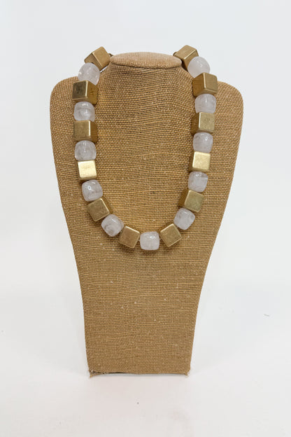 A chunky beaded necklace with alternating matte gold square beads and translucent white-gray round beads, displayed on a tan fabric-covered jewelry bust against a white background.