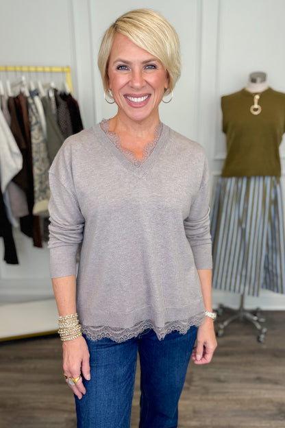 Front view of woman wearing Swagger Boutique’s Mocha Lace Lined V-Neck Sweater  with jeans with clothing hanging in the background.