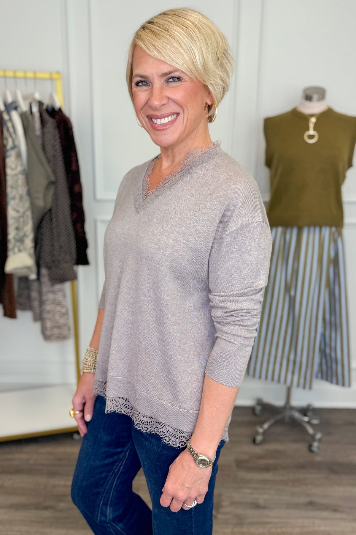 Side view of woman wearing Swagger Boutique’s Mocha Lace Lined V-Neck Sweater  with jeans with clothing hanging in the background.