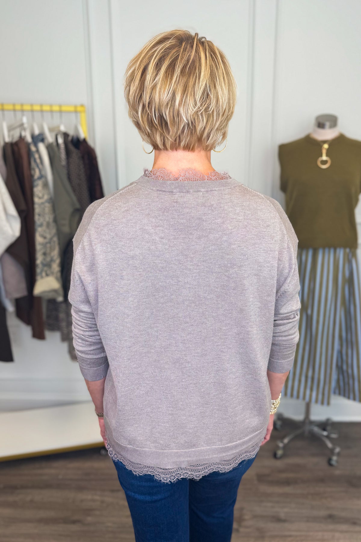 Back view of woman wearing Swagger Boutique’s Mocha Lace Lined V-Neck Sweater  with jeans with clothing hanging in the background.