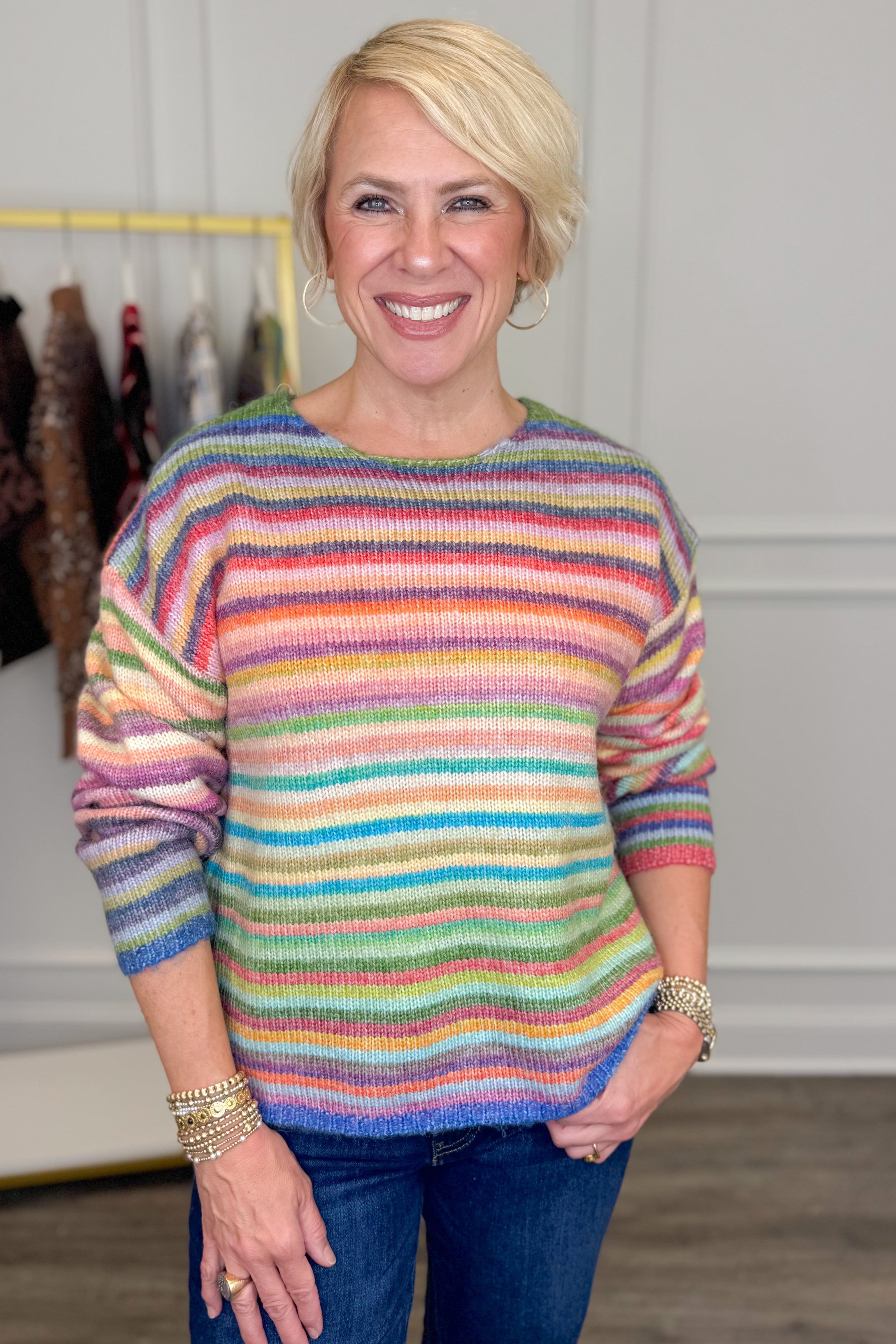 Front view of woman wearing Swagger Boutique’s Multi Striped Sweater with jeans with clothing hanging in the background.