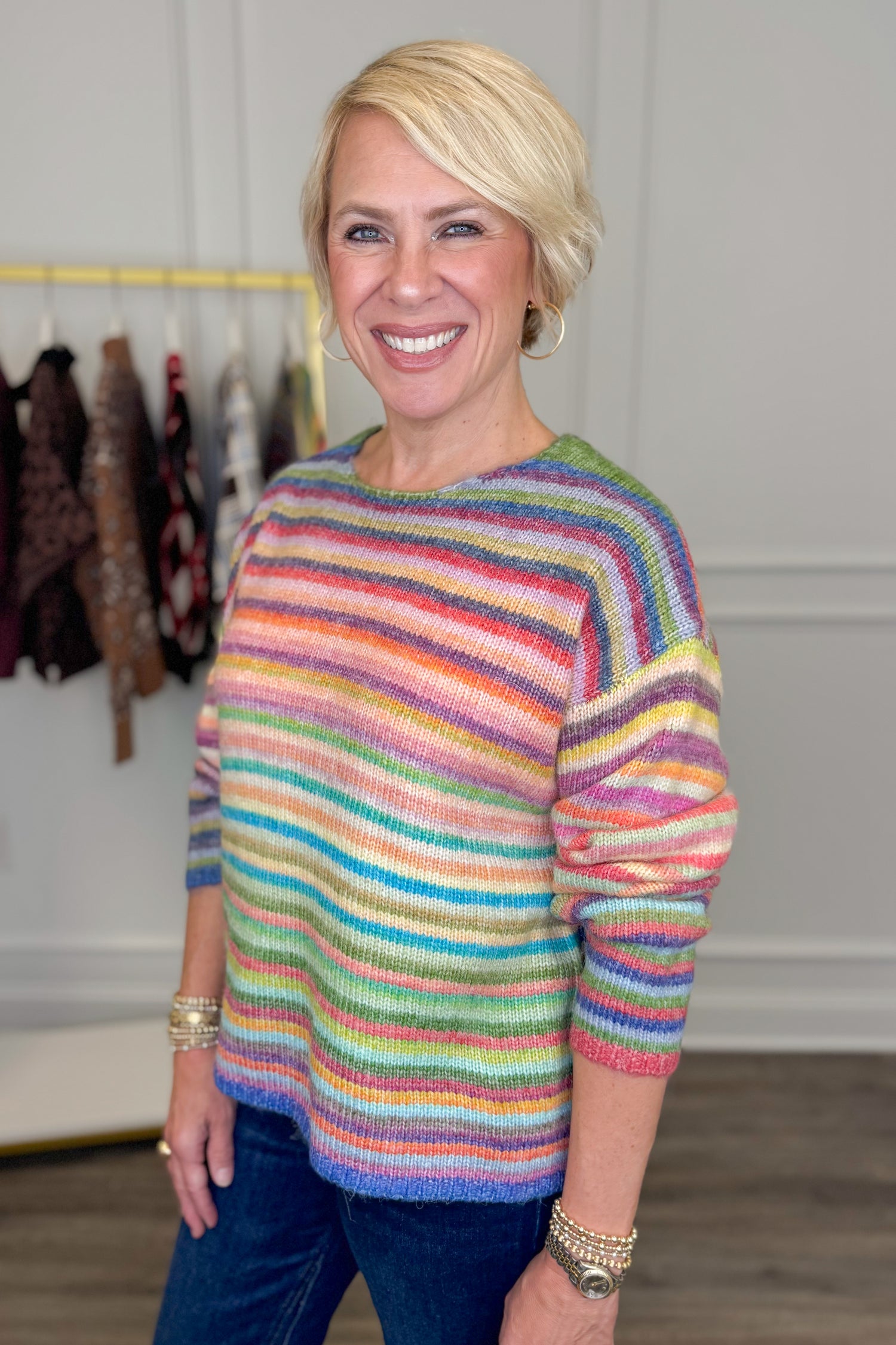 Side view of woman wearing Swagger Boutique’s Multi Striped Sweater with jeans with clothing hanging in the background.