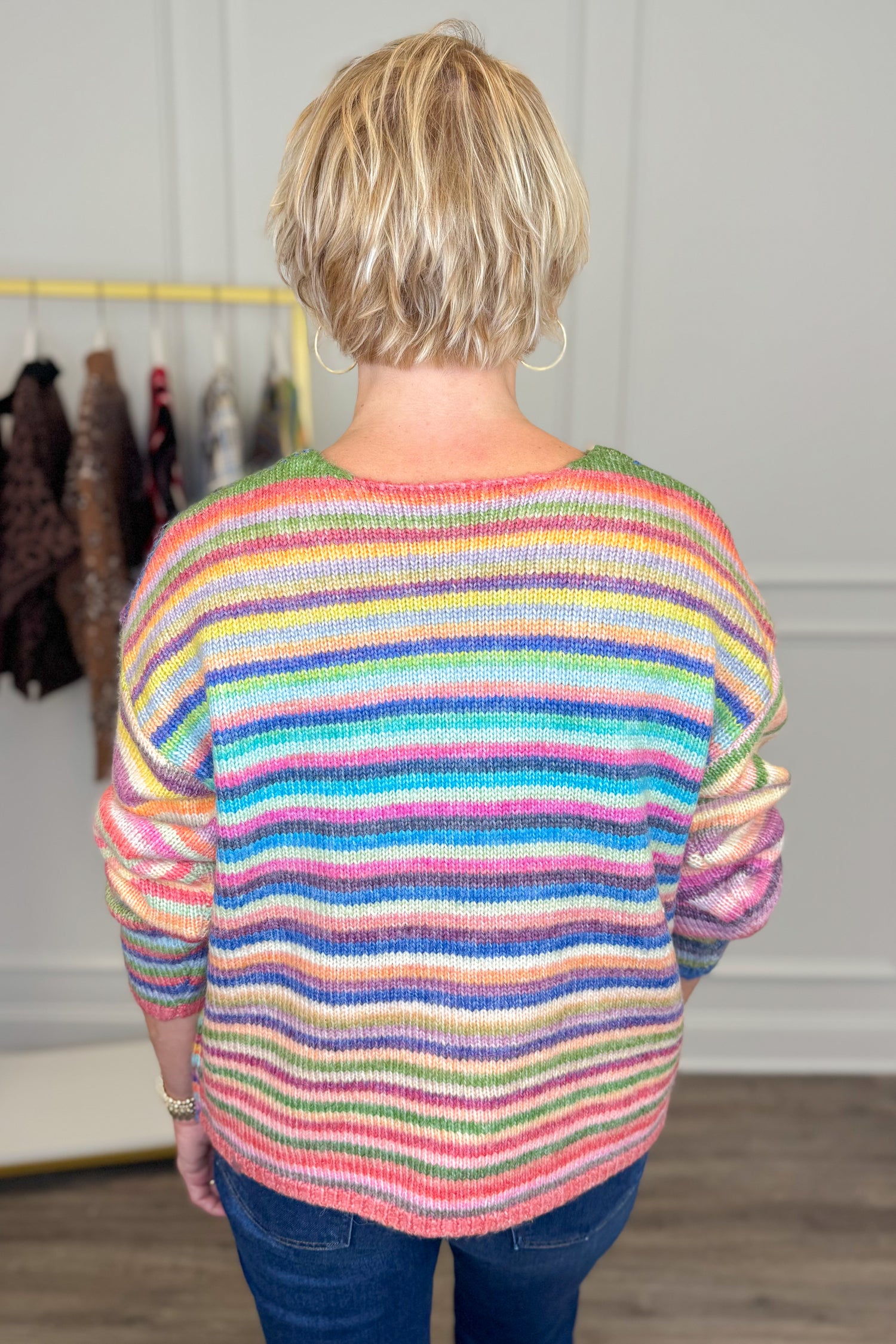 Back view of woman wearing Swagger Boutique’s Multi Striped Sweater with jeans with clothing hanging in the background.