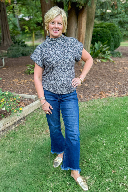 Front view of woman wearing Grey Leopard Short Sleeve Fuzzy Top and blue jeans outside with greenery and trees.