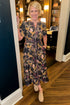 Front view of woman wearing Swagger Boutique’s Navy Animal Print Midi Dress with clogs standing outside of a den.