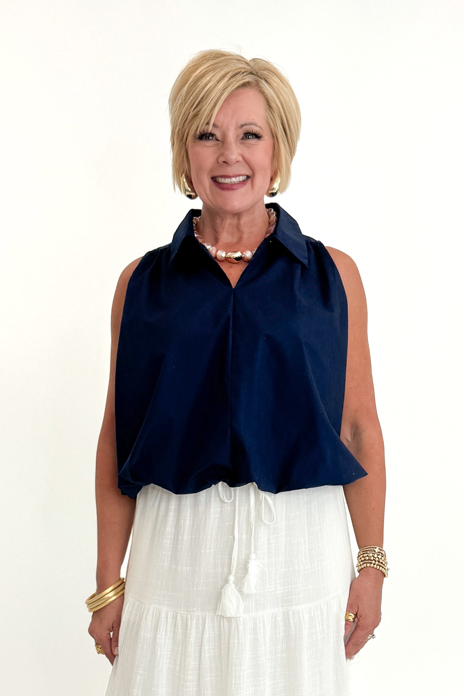 Front view of woman wearing Swagger Boutique’s Navy Bubble Hem Collared Sleeveless Top with Just A Consideration White Flowy Tiered Cotton Skirt against white studio background.