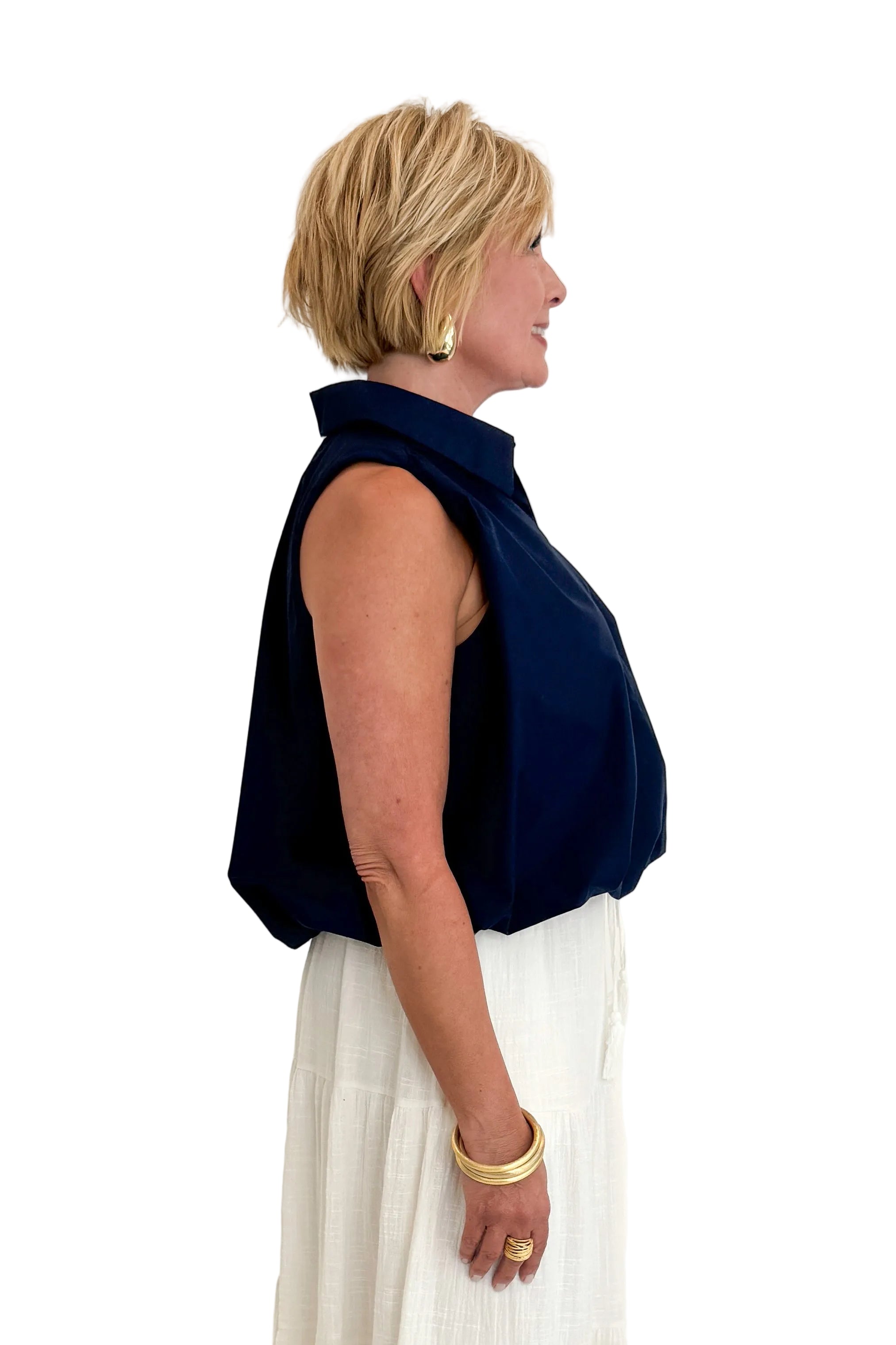 Side view of woman wearing Swagger Boutique’s Navy Bubble Hem Collared Sleeveless Top with Just A Consideration White Flowy Tiered Cotton Skirt against white studio background.
