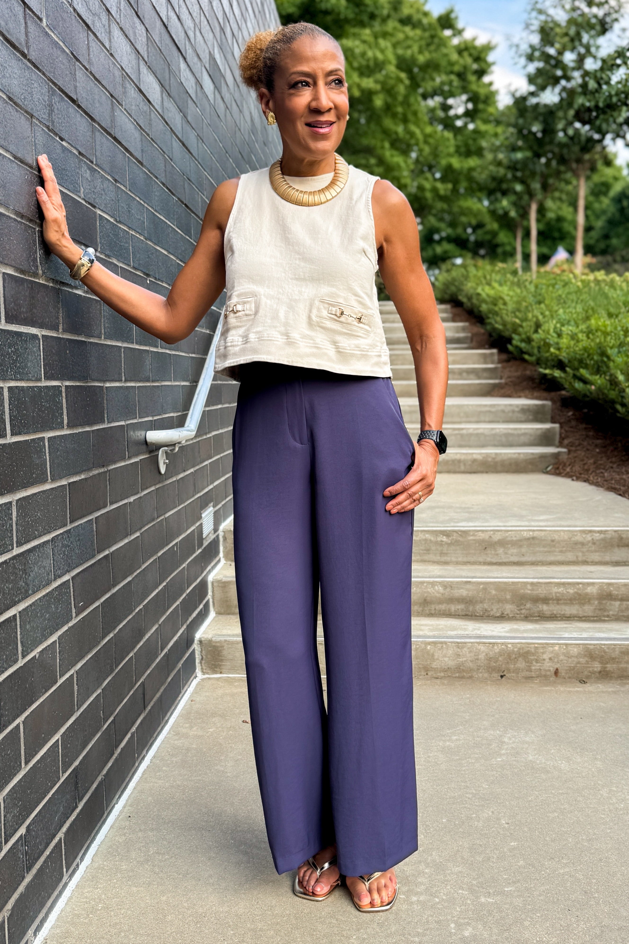 Front view of woman wearing Swagger Boutique’s Ecru Horsebit Buckle Tank by Entro with Navy High Waisted Trousers by Skies Are Blue standing against a brick wall.