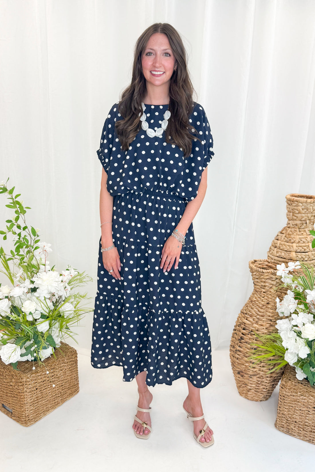 Front view of woman wearing Swagger Boutique’s Navy Polka Dot Bubble Hem Top by Entro with matching Navy Polka Dot Tiered Skirt by Entro against decorative background.