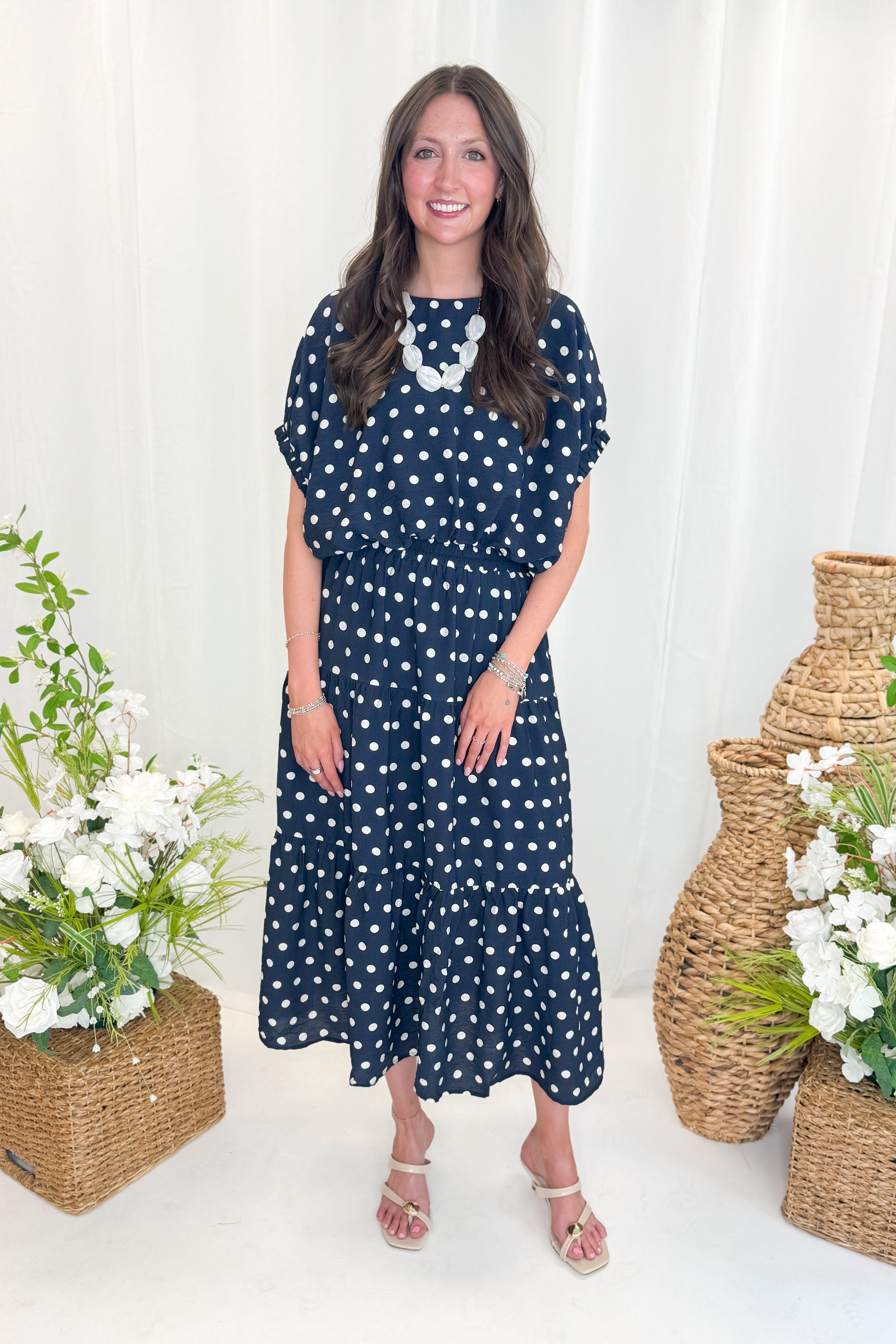 Front view of woman wearing Swagger Boutique’s Navy Polka Dot Bubble Hem Top by Entro with matching Navy Polka Dot Tiered Skirt by Entro against decorative background.