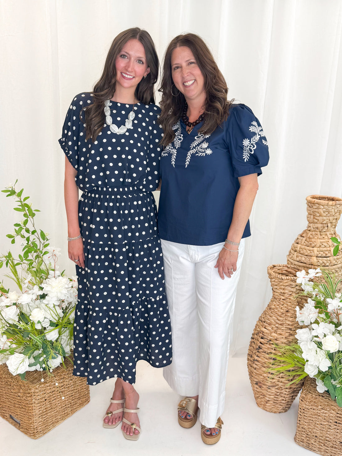 Front view of woman wearing Swagger Boutique’s Navy Polka Dot Bubble Hem Top by Entro with matching Navy Polka Dot Tiered Skirt by Entro and woman wearing Navy &amp; White Embroidered V-Neck Top against decorative background.