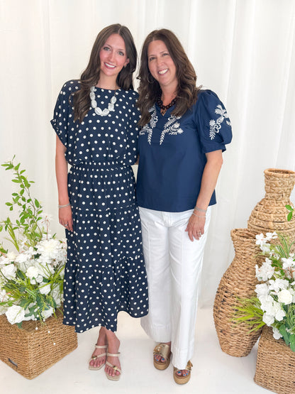 Front view of woman wearing Swagger Boutique’s Navy Polka Dot Bubble Hem Top by Entro with matching Navy Polka Dot Tiered Skirt by Entro and woman wearing Navy &amp; White Embroidered V-Neck Top against decorative background.