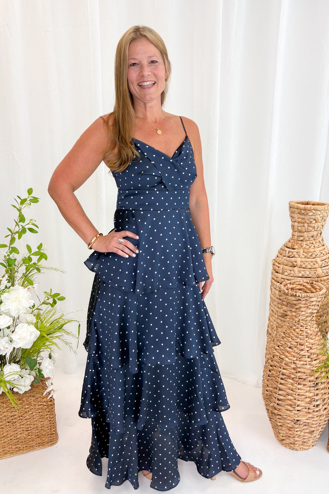 Front view of woman wearing Swagger Boutique’s Navy Polka Dot Tiered Maxi Dress against decorative background.