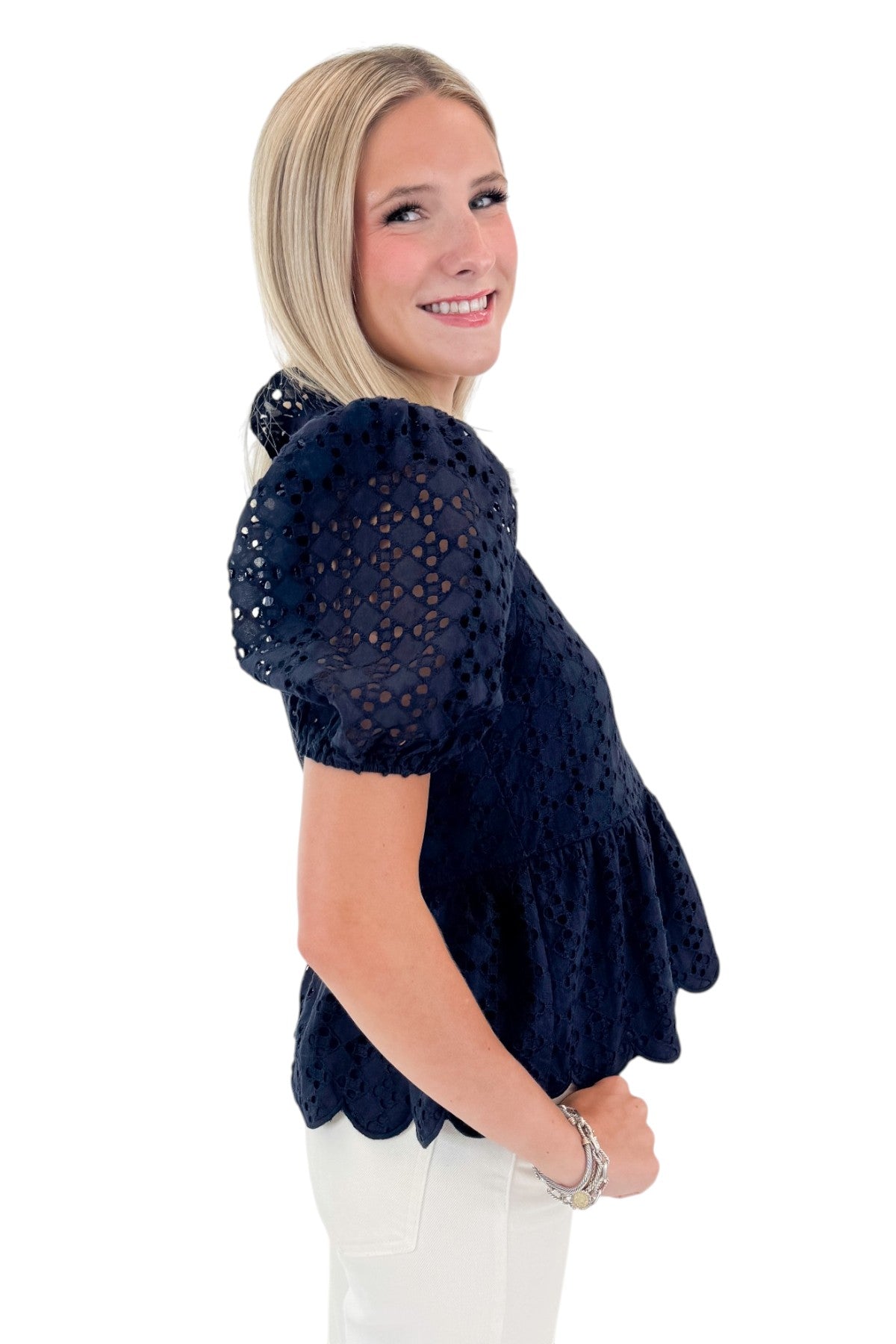 Side view of woman wearing Navy Short Sleeve Peplum Eyelet Top with jeans against white studio background.