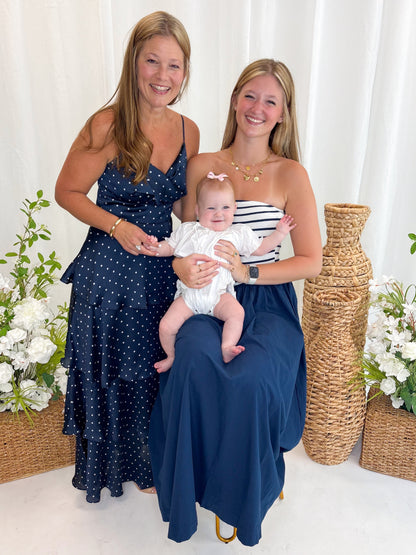 Front view of woman wearing Swagger Boutique’s Navy Striped Maxi Dress and Navy Polka Dot Tiered Maxi Dress with sandals against white decorative background.
