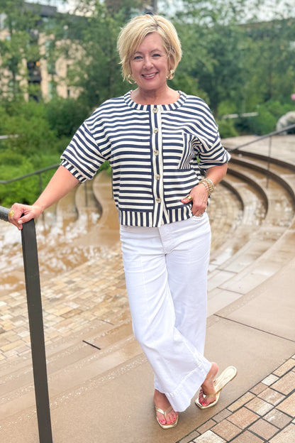 Front view of woman wearing Swagger Boutique’s Navy Striped Short Sleeve Button Up Top with jeans leaning against a railing outside.
