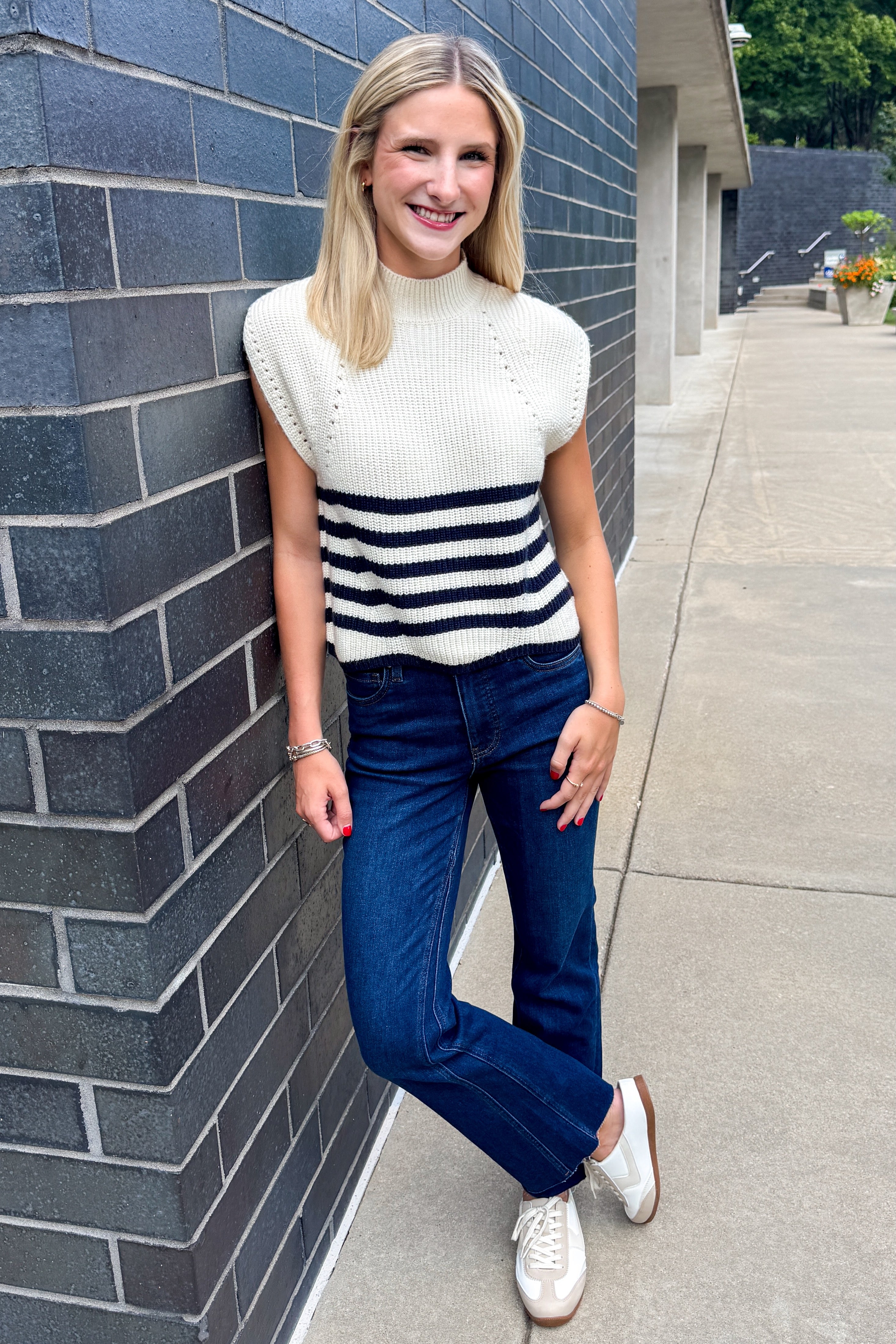 Front view of woman wearing Swagger Boutique’s Navy Striped Turtleneck Sleeveless Sweater with jeans standing outside against a brick wall.